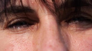 Gözünü kırpıp dışarıdaki kameraya bakan bir kadının yorgun mavi gözleri. Güneşin parlak ışıklarıyla aydınlatan yetişkin bir kadının yüzü. Hanımefendinin üzgün yüz ifadesi. Yavaşla.