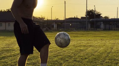 Genç adam gün batımında stadyumda futbol topuyla hokkabazlık yapıyor. Yeşil sahada profesyonel futbolcu topa vuruyor. Sporcu arka planda güneş ışığıyla çayırda numaralar yapıyor. Serbest stil futbol.