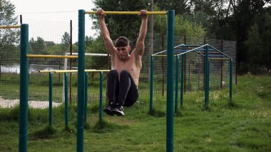 Atletik adam karın kaslarını eğitir spor sahasında yatay barda bacak kaldırır. Genç sporcu, barfiks çekme çubuğunda egzersiz yapıyor. Doğada spor yapan sporcu. Sağlıklı yaşam tarzı.