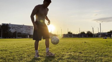 Genç adam gün batımında stadyumda futbol topuyla hokkabazlık yapıyor. Yeşil sahada profesyonel futbolcu topa vuruyor. Sporcu arka planda güneş ışığıyla çayırda numaralar yapıyor. Serbest stil futbol.