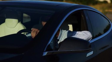 Modern elektrikli arabanın ön camından bakan yakışıklı başarılı bir girişimci. Yenilikçi elektrikli aracında gülümseyen genç iş adamının portresi. Yavaş çekim