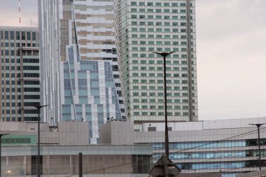 Yüksek gökdelenleri, ofis kuleleri, yansıtıcı cam pencereleri ve bulutlu gökyüzü altında geometrik cepheleri olan çağdaş şehir silueti kentsel gelişimi ve modern mimariyi gözler önüne seriyor.