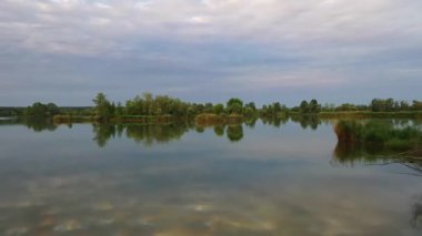 Ağaçlar arasında kıyıdan uçan ve gölün üzerinde yavaşça yükselen bir hava aracı videosu. Pürüzsüz ileri hareket küçük adaları, sudaki yansımaları ve geniş manzaralı manzarayı ortaya çıkarır..