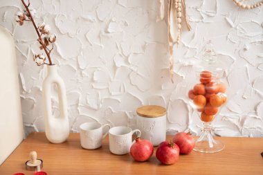 A cozy display of pomegranates and ceramic mugs on a warm wooden table against a textured white wall clipart