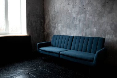 Minimalist, gri duvarları ve koyu renkli zemini olan, pencerenin yanına kurulmuş rahat, mavi bir kanepe, huzurlu bir atmosfer yaratıyor.