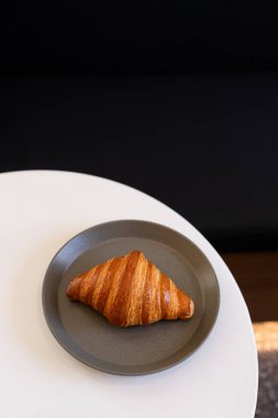 Fırından yeni çıkmış bir kruvasan, kahvaltı saatlerinde beyaz bir masada, sıcak bir yerde, gri bir tabakta oturuyor.
