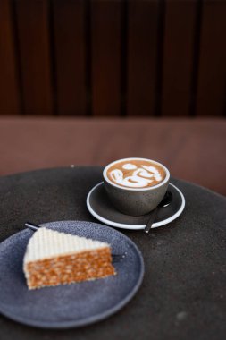 Lezzetli kahve, sanatsal latte tasarımı ve bir dilim karamel keki, rahat bir kafede bir masada servis ediliyor.