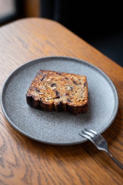 Gri tabakta taze pişmiş muz ekmeği, rahat bir kafede, ahşap masada çatalla servis edilir.