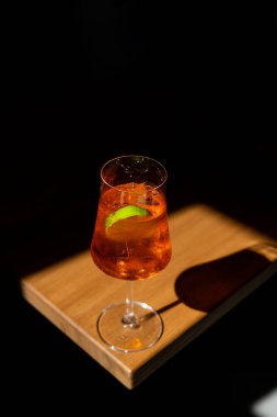 Narenciye kokteyli, şeffaf bardakta servis edilir. Ahşap bir masada koyu bir arka planda limon dilimi ile servis edilir.