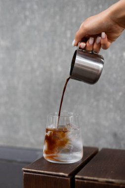 Kaliteli bir bardakta buzun üzerine zengin bir kahve karışımı dökmek ahşap bir masada modern bir kafeterya ortamı sergiliyor.