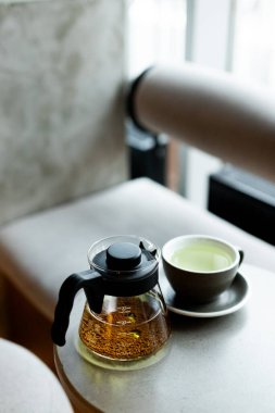 A cozy tea moment with a glass teapot and cup placed on a soft surface by a window in a serene setting clipart