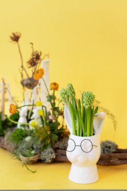 Kaprisli bir beyaz tavşan yetiştiricisi açık sarı bir arka plana karşı canlı yeşillik tutar, kapalı alanda eğlenceli bir bahçe düzenlemesi sergiler.