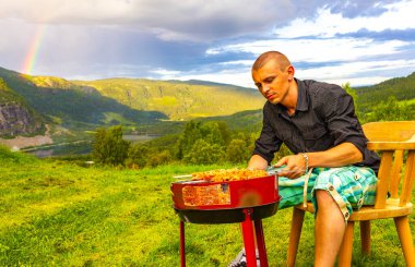 Genç erkek çocuk İskandinavya 'daki Grindaheim Vang i Valdres Innlandet Norveç' te dağ manzarası ve gökkuşağıyla ızgara yapıyor..