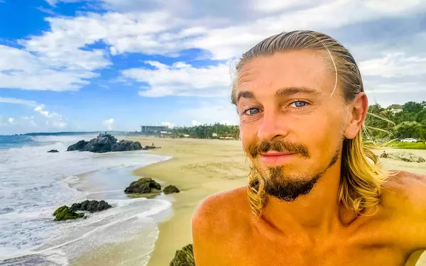 Male tourist traveler man makes selfie with beautiful rocks cliffs stones boulders and huge big surfer waves and natural panorama view on the beach in Puerto Escondido Oaxaca Mexico.