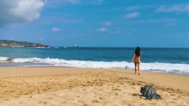 Seksi bayan turist ve renkli bikinili gezgin kız ve dövmeleri olan Mazunte Oaxaca Meksika 'da kum dalgaları ve dağ uçurumları olan tropik sahillerde yürüyor ve yüzüyor..