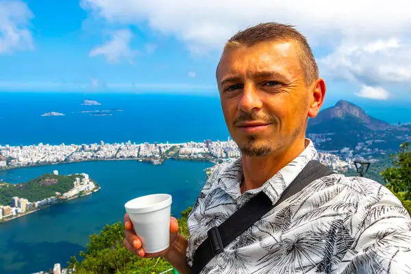 Kahve fincanıyla yakışıklı erkek turist ve Brezilya 'da Rio de Janeiro Panorama City Skyline Plaj Sahili Deniz Dağları Tropik Orman ve Mavi Bulutlu Gök manzarası.