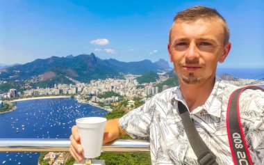 Kahve fincanıyla yakışıklı erkek turist ve Brezilya 'da Rio de Janeiro Panorama City Skyline Plaj Sahili Deniz Dağları Tropik Orman ve Mavi Bulutlu Gök manzarası.