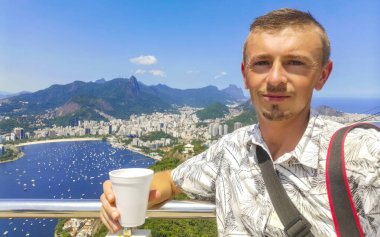 Kahve fincanıyla yakışıklı erkek turist ve Brezilya 'da Rio de Janeiro Panorama City Skyline Plaj Sahili Deniz Dağları Tropik Orman ve Mavi Bulutlu Gök manzarası.