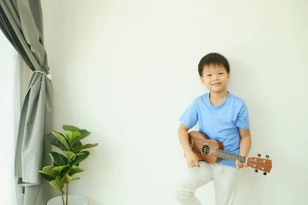 Cute Asian happy smiling 7 years old school boy looking at camera  having fun playing ukulele in the living room at home, Music and hobby for kids concept