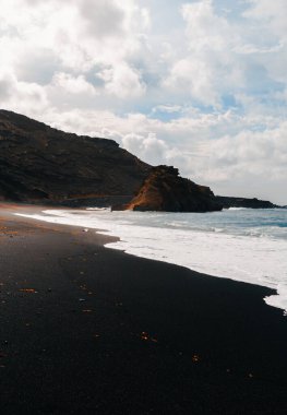 Lanzarote (Kanarya Adaları) volkanik adasındaki El Golfo plajında mavi okyanusta güçlü dalgalarla çekilmiş dikey bir fotoğraf. Arka planda bulutlu mavi gökyüzü ile vahşi siyah kumsalda uçurum.