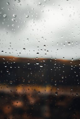 Arka planda bulutlu gökyüzü olan penceredeki yağmur damlalarının resmini kapat. Yağmur damlalarının dikey görüntüsü pencereye düşer ve arka planda yağmurlu bir günde yağmur yağar.