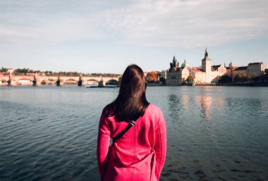 Prag 'da (Çek Cumhuriyeti) Vltava nehrinin yanında duran mor paltolu kadın gezgin gün batımında Charles Köprüsü' ne bakıyor. Şehirdeki kadın turistin geniş açı çekimi.