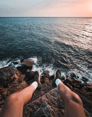 POV Manzaralı - erkek turist (erkek) okyanusun üzerindeki uçurumun kenarında oturuyor. Denizin üstünde bacakları olan bir adam ufukta inanılmaz bir gün batımı izliyor..