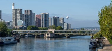 Paris 'teki nehir kıyısında gezinti güvertesi, demirli gemiler, sıcak hava balonu ve modern ofis binaları.