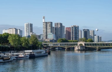 Paris 'teki nehir kıyısında gezinti güvertesi, demirli gemiler, sıcak hava balonu ve modern ofis binaları.