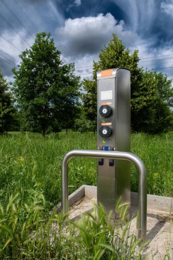 Ağaçların ve elektrik kablolarının önündeki yeşil çayırdaki elektrikli arabalar için şarj istasyonu.