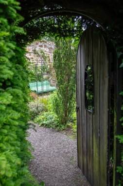 Eski ahşap kapı açık duruyor ve dinlenmek için banklı huzurlu bahçeye erişim sağlıyor. 