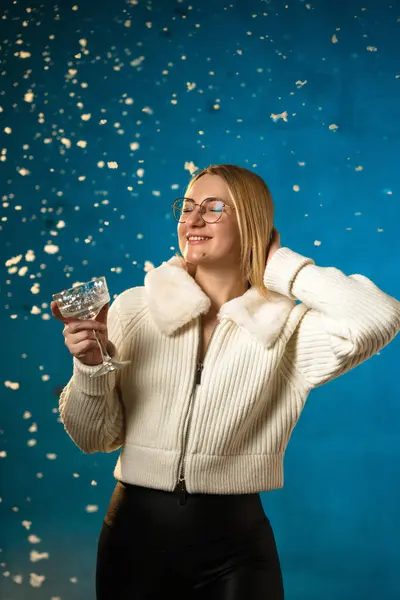 青い背景に白い毛皮のセーターのブロンドかわいい慎重な女性 偽の雪が彼女の上を流れています 彼女は幸せで クリスマスと新年を祝います スタジオポートレート ロイヤリティフリーのストック画像