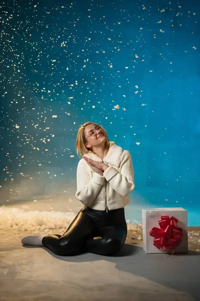 穿着蓝色背景的白色毛皮毛衣的金发美女 假雪在她身上飞舞 她很高兴 坐在礼物上 庆祝圣诞节和新年 工作室肖像 图库图片