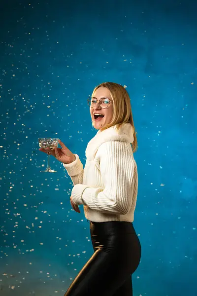 穿着蓝色背景的白色毛皮毛衣的金发美女 假雪在她身上飞舞 她很高兴 庆祝圣诞节和新年 工作室肖像 免版税图库照片