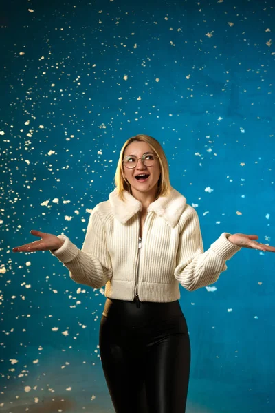 穿着蓝色背景的白色毛皮毛衣的金发美女 假雪在她身上飞舞 她很高兴 庆祝圣诞节和新年 工作室肖像 免版税图库图片