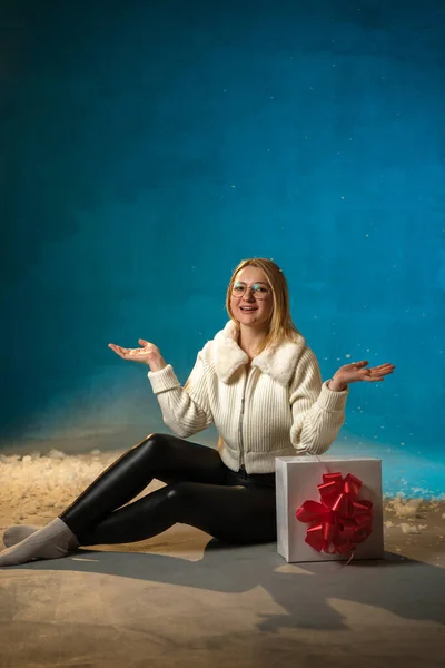 青い背景に白い毛皮のセーターのブロンドかわいい慎重な女性 偽の雪が彼女の上を流れています 彼女は幸せで プレゼントに座って クリスマスと新年を祝います スタジオポートレート ストック画像