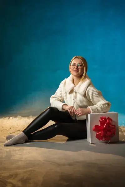青い背景に白い毛皮のセーターのブロンドかわいい慎重な女性 偽の雪が彼女の上を流れています 彼女は幸せで プレゼントに座って クリスマスと新年を祝います スタジオポートレート ロイヤリティフリーのストック写真