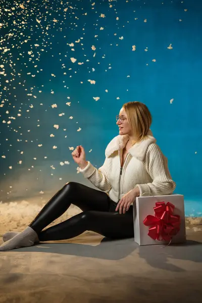 穿着蓝色背景的白色毛皮毛衣的金发美女 假雪在她身上飞舞 她很高兴 坐在礼物上 庆祝圣诞节和新年 工作室肖像 免版税图库照片