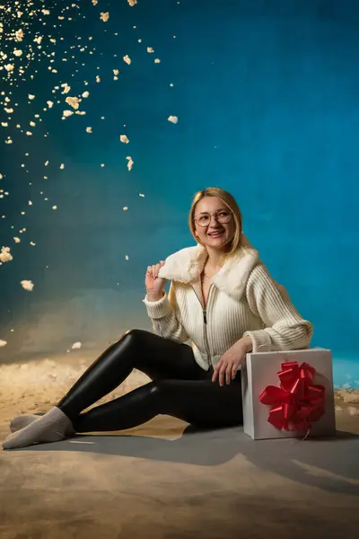 穿着蓝色背景的白色毛皮毛衣的金发美女 假雪在她身上飞舞 她很高兴 坐在礼物上 庆祝圣诞节和新年 工作室肖像 免版税图库图片