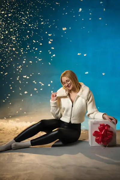 青い背景に白い毛皮のセーターのブロンドかわいい慎重な女性 偽の雪が彼女の上を流れています 彼女は幸せで プレゼントに座って クリスマスと新年を祝います スタジオポートレート ストックフォト