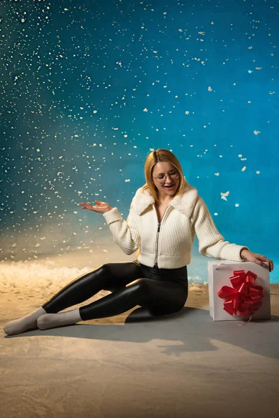 穿着蓝色背景的白色毛皮毛衣的金发美女 假雪在她身上飞舞 她很高兴 坐在礼物上 庆祝圣诞节和新年 工作室肖像 图库照片