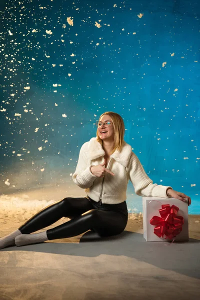 青い背景に白い毛皮のセーターのブロンドかわいい慎重な女性 偽の雪が彼女の上を流れています 彼女は幸せで プレゼントに座って クリスマスと新年を祝います スタジオポートレート ストック写真