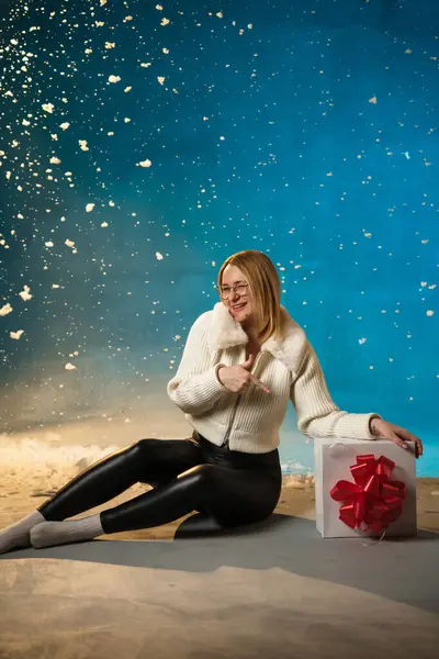 青い背景に白い毛皮のセーターのブロンドかわいい慎重な女性 偽の雪が彼女の上を流れています 彼女は幸せで プレゼントに座って クリスマスと新年を祝います スタジオポートレート ストック画像