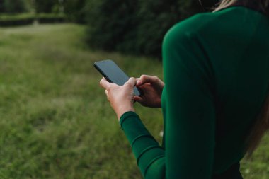 Parkta güneş ışığıyla yoga. Lotus pozisyonunda genç bir kadın yeşil çimlerin üzerinde oturuyor ve cep telefonu kullanıyor. Dijital dönüşüm kavramı.