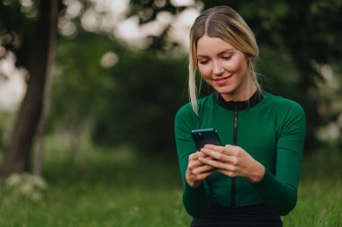 Parkta güneş ışığıyla yoga. Lotus pozisyonunda genç bir kadın yeşil çimlerin üzerinde oturuyor ve cep telefonu kullanıyor. Dijital dönüşüm kavramı.