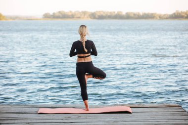 Genç ve güzel bir kız, nehrin kıyısında sakin ve sıcak bir akşamda yoga yapıyor..