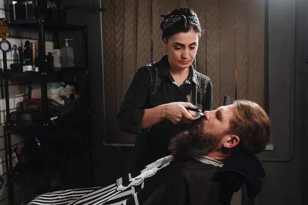 Young woman barber making haircut of bearded man in barbershop. Self-care, masculine beauty. Hair care service concept