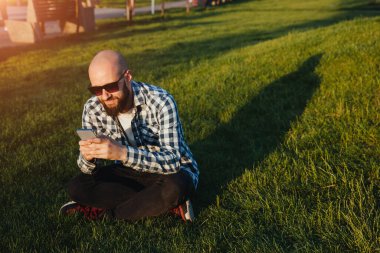 A young man sits on the green grass in the park and uses social networks using a mobile phone.