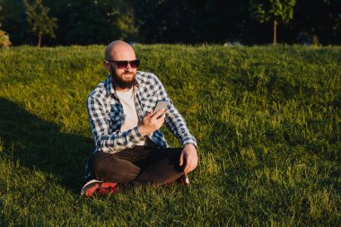 A young man sits on the green grass in the park and uses social networks using a mobile phone.