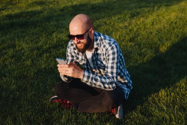 A young man sits on the green grass in the park and uses social networks using a mobile phone.